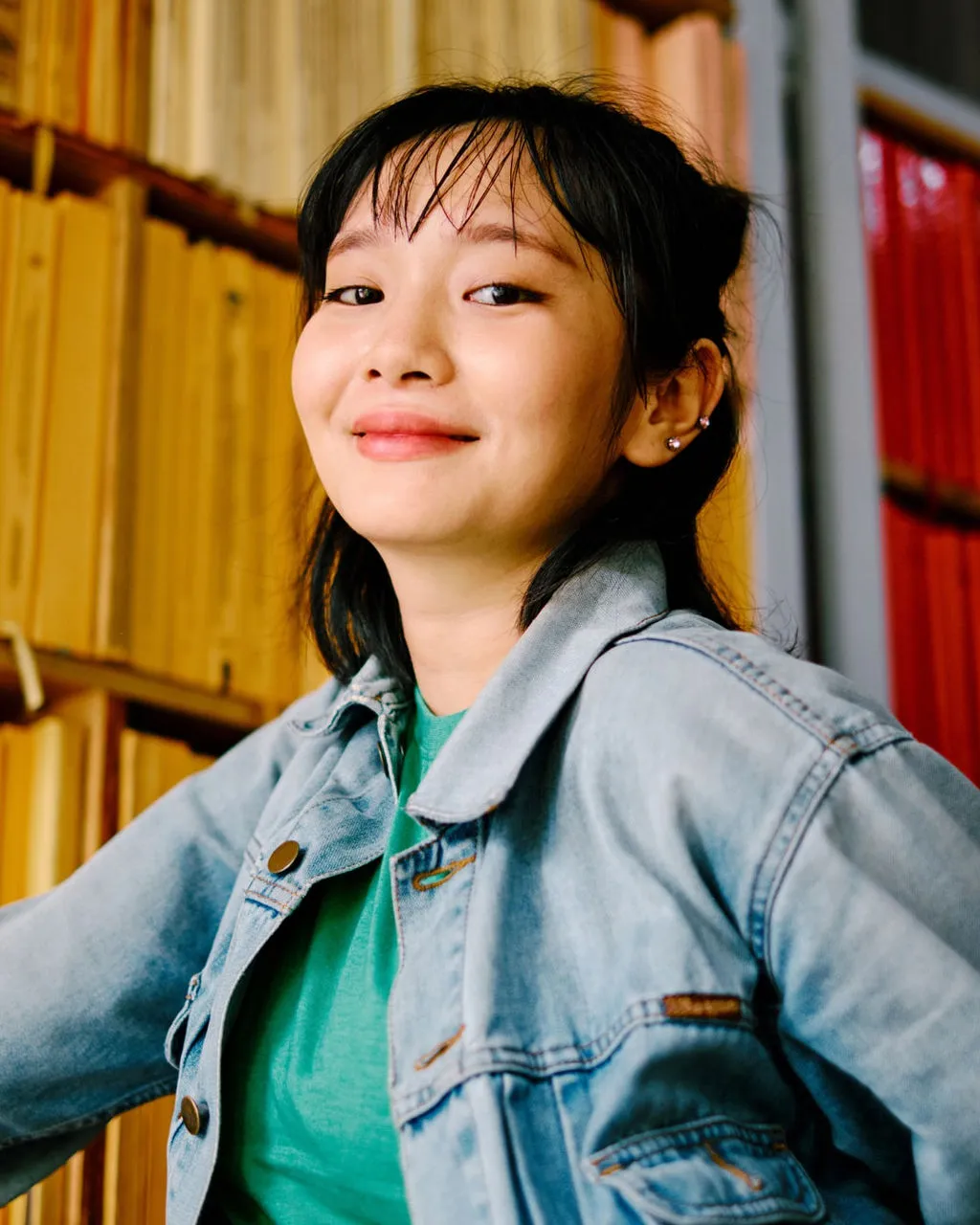 A young woman in a denim jacket smiles proudly in front of a shelf full of books. 