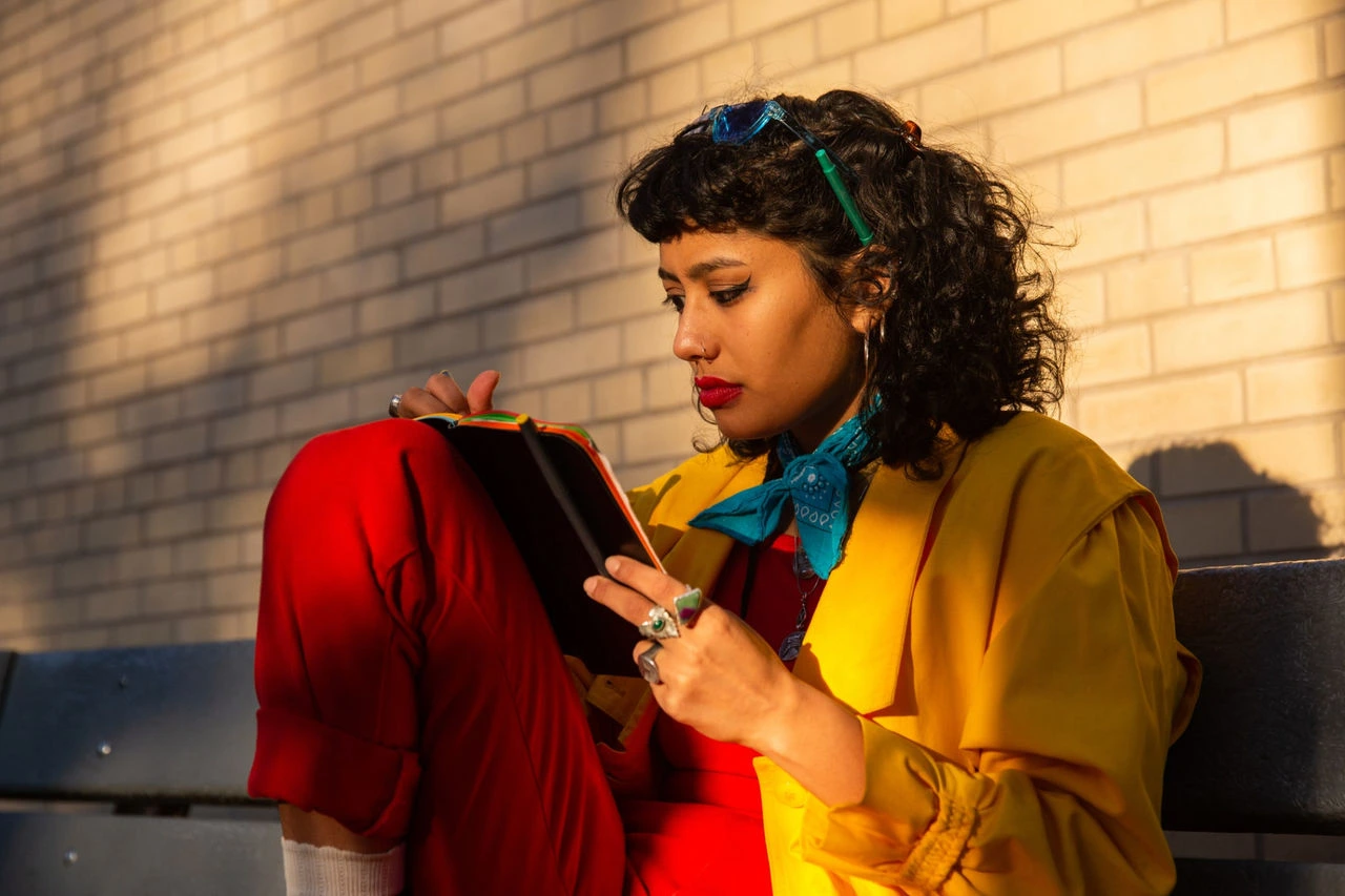 A woman in a brightly colored outfit leans against a brick wall with her tablet. She is focused on drawing something on the screen. 