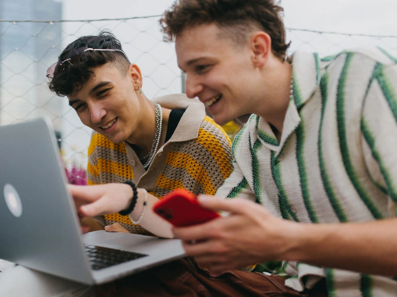 Two young men are smiling and looking at a laptop with interest. One is holding a smart phone and pointing at the screen while the second leans closer.